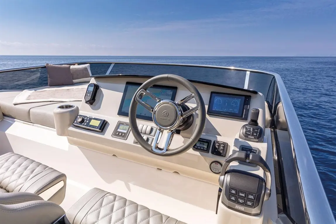 Boat cockpit view with steering wheel, control screens, and plush seating, overlooking calm water under a blue sky.